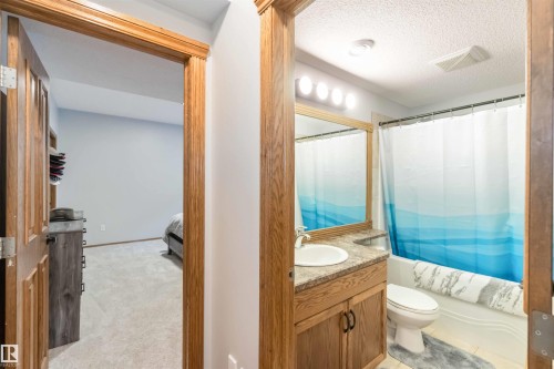The property features a bathroom with a vanity, mirror, toilet, and a bathtub with a shower curtain, alongside a carpeted room with light colored walls - 61 Linkside Blvd, Spruce Grove, AB 