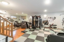 This spacious lower level features a dedicated workout area with checkered floor tiling and recessed lighting, and a separate living area with wood-style flooring and a patterned area rug - 