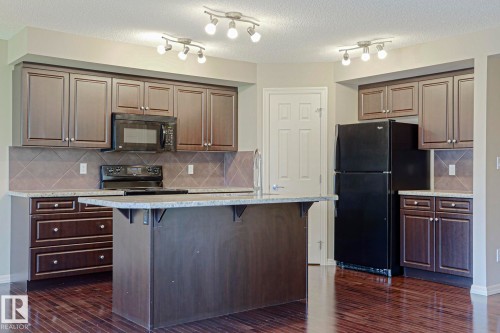 Open-concept kitchen featuring dark wood cabinetry, granite countertops, tile backsplash, a central island, and black appliances - 6215 60 Street, Beaumont, AB - Indoor Photo Showing Kitchen