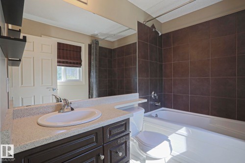 Bathroom featuring a dark wood-finish vanity with a light speckled countertop, an integrated sink, a full-width mirror, a tub/shower combination with dark square wall tiles, and chrome fixtures - 6215 60 Street, Beaumont, AB - Indoor Photo Showing Bathroom