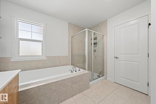 Bathroom featuring a built-in tub, separate glass-enclosed shower, light-toned tile flooring, and a window with frosted glass - 1931 Westerra Lane, Stony Plain, AB 
