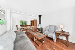 Open-concept living area featuring rich wood-finish flooring, a freestanding wood-burning stove, and a dining space with a window overlooking exterior greenery - 