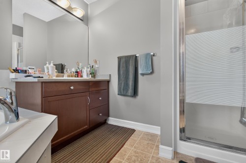 Bathroom featuring a wood-finish vanity with a light-toned countertop, an integrated mirror with overhead lighting, a glass-enclosed shower, and neutral-toned floor tiling - 547 Leger Way, Edmonton, AB 