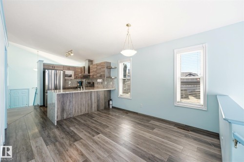 Open-concept kitchen and dining area featuring wood-finish flooring, a kitchen island with stone countertop, stainless steel appliances, and two windows with blinds - 14908 137 Street, Edmonton, AB 
