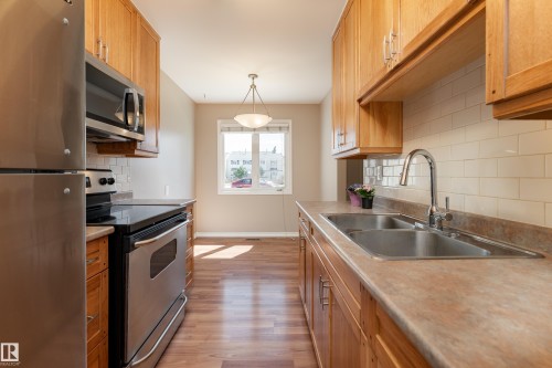 Galley kitchen featuring wood-finish cabinetry, stainless steel appliances, a double basin sink, and a subway tile backsplash - 16B Callingwood Court, Edmonton, AB 