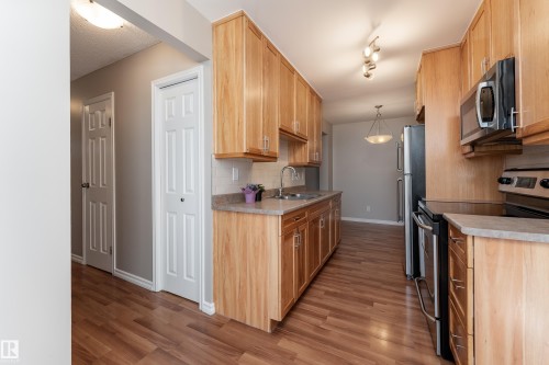 Galley kitchen featuring wood-finish cabinetry, stainless steel appliances, stone-look countertops, and wood-finish flooring - 16B Callingwood Court, Edmonton, AB 