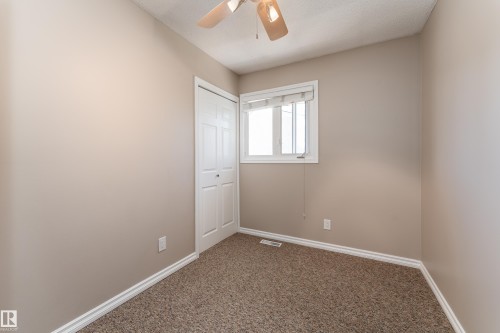 Neutral-toned room featuring a white-framed window with blinds, bi-fold closet doors, and textured carpet flooring - 16B Callingwood Court, Edmonton, AB 