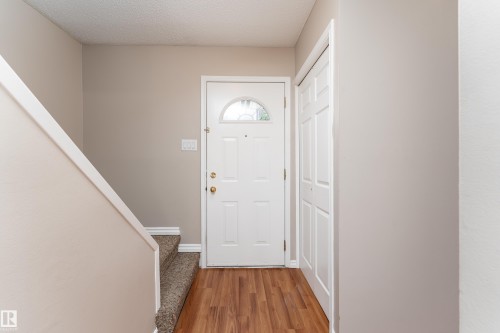 Entryway featuring wood-finish flooring, a white paneled door with an arched glass insert, a white bi-fold closet door, and a carpeted staircase with a white handrail - 16B Callingwood Court, Edmonton, AB 