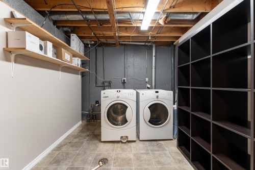 Dedicated laundry area featuring a front-loading washer and dryer, tile-finish flooring, exposed ceiling joists, and built-in shelving - 16B Callingwood Court, Edmonton, AB 
