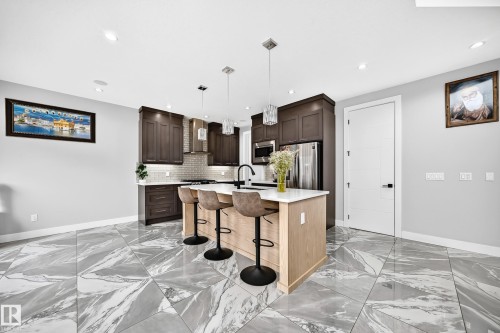 Kitchen featuring a light wood-finish island with a white countertop and three pendant lights, dark wood-finish cabinetry, a stainless steel range hood, and large format grey and white patterned floor tiles - 8710 Mayday Lane, Edmonton, AB 
