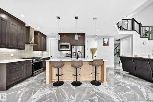 Open-concept kitchen featuring an island with seating, stainless steel appliances, dark wood cabinetry, a subway tile backsplash, and large format grey marble-effect floor tiles - 8710 Mayday Lane, Edmonton, AB 