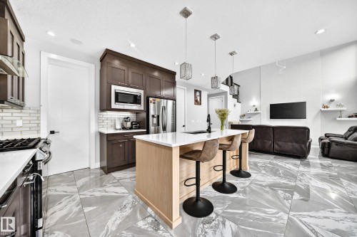 Open-concept kitchen and living area featuring a central island with seating, stainless steel appliances, dark wood cabinetry, and large format marble-patterned floor tiles - 8710 Mayday Lane, Edmonton, AB 