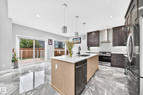 Modern kitchen featuring a light wood-finish island with a white countertop and undermount sink, dark wood-finish cabinetry, stainless steel appliances, and a white subway tile backsplash - 8710 Mayday Lane, Edmonton, AB 