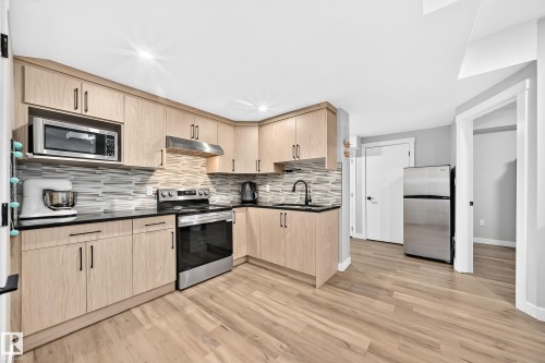 Kitchen featuring light wood-finish cabinetry, black countertops, and a mosaic tile backsplash - 8710 Mayday Lane, Edmonton, AB 