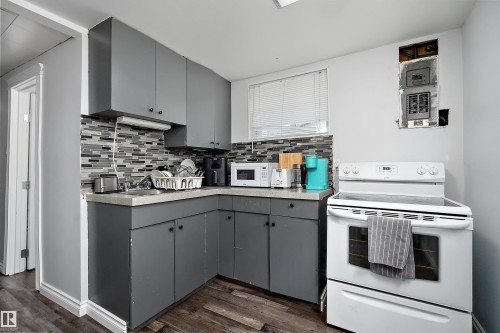 Compact kitchen featuring grey cabinetry, a mosaic tile backsplash, a white electric range, and wood-finish flooring - 5516 47A Avenue, Wetaskiwin, AB - Indoor Photo Showing Kitchen