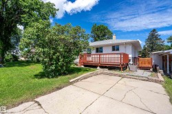 Spacious backyard featuring a large wood-finish deck with railings, mature trees, and a concrete patio area with brick retaining walls - 