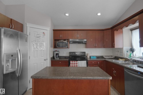 Kitchen featuring wood-finish cabinetry, a freestanding central island with a granite-style countertop, a stainless steel refrigerator, and a double basin sink - 4816 155 Ave, Edmonton, AB 
