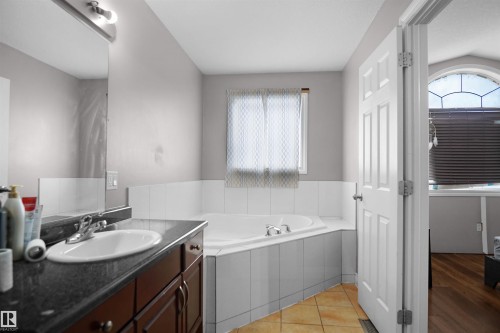 Bathroom featuring a corner soaking tub with white tile surround, a single vanity with a dark stone countertop, a rectangular mirror, and beige floor tiles - 4816 155 Ave, Edmonton, AB 