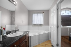 Bathroom featuring a corner soaking tub with white tile surround, a single vanity with a dark stone countertop, a rectangular mirror, and beige floor tiles - 