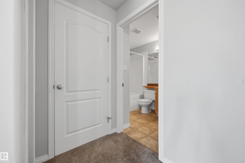 Hallway to the storage unit, bathroom to the right, and secondary bedroom to the left. Featuring neutral-toned carpeting and light gray wall paint - 201 3425 19 Street Nw, Edmonton, AB 