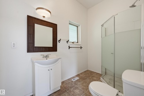 Bathroom featuring a white vanity with integrated sink, a framed mirror, a frosted glass shower enclosure, a toilet, and neutral-toned tile flooring - 11450 71 Street Nw, Edmonton, AB 