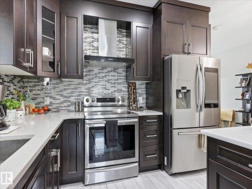 Modern kitchen featuring dark wood cabinetry, stainless steel appliances, a multi-tone tile backsplash, light-toned countertops, and light wood-finish flooring - 12820 68 Street, Edmonton, AB - Indoor Photo Showing Kitchen With Upgraded Kitchen