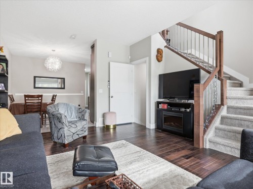 Open-concept living space featuring wood-finish flooring, a built-in electric fireplace, a carpeted staircase with wood and metal railing, and a dining area with a modern chandelier - 12820 68 Street, Edmonton, AB - Indoor Photo Showing Living Room