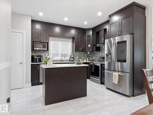 Modern kitchen featuring dark wood cabinetry, a central island with a light-toned countertop, stainless steel appliances, and a patterned tile backsplash - 12820 68 Street, Edmonton, AB - Indoor Photo Showing Kitchen With Upgraded Kitchen