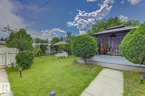 Expansive backyard featuring a screened gazebo on a raised deck, a manicured lawn, mature shrubs, a concrete pathway, and a white picket fence - 15707 92 Avenue, Edmonton, AB 