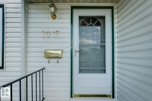 White siding exterior featuring a storm door with an arched window, brass house numbers, a coordinating brass mailbox, and a black metal handrail - 3015 131 Avenue, Edmonton, AB 