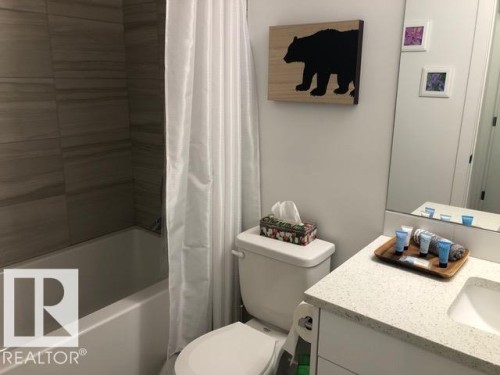 Bathroom featuring a shower-over-tub combination with horizontal tile surround, a white vanity with an integrated rectangular sink, a stone-finish countertop, and a built-in mirror - 216 11503 76 Avenue, Edmonton, AB 