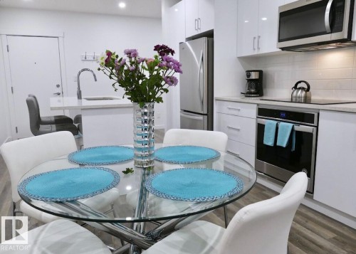 Kitchen featuring wood-finish flooring, white cabinetry, integrated stainless steel appliances, and a central island with a sink and seating - 216 11503 76 Avenue, Edmonton, AB 