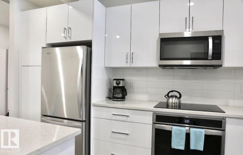 Modern kitchen featuring glossy white cabinetry, stainless steel appliances, a white tile backsplash, and light-toned countertops - 216 11503 76 Avenue, Edmonton, AB 