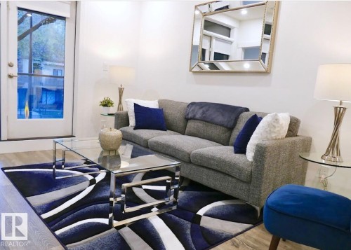 Spacious living area featuring light wood-finish flooring, a full-light glass panel door, white walls, and a decorative wall mirror - 216 11503 76 Avenue, Edmonton, AB 