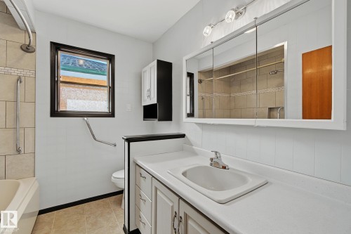 Bathroom featuring a vanity with an integrated sink, tri-fold medicine cabinet with overhead lighting, a tub/shower combination with tiled surround, and a wall-mounted storage cabinet - 9545 65 Avenue, Edmonton, AB 