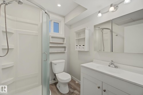 Bathroom featuring a standing shower with a curved glass enclosure, a white vanity with an integrated sink, a mirrored medicine cabinet, a window, and wood-finish flooring - 9545 65 Avenue, Edmonton, AB 