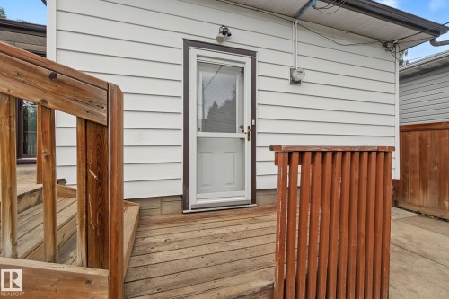Decked exterior with white siding, showcasing wood-finish steps and railing, a storm door entry, and a partial privacy fence - 9545 65 Avenue, Edmonton, AB 