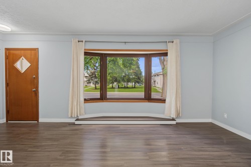 Bay window with dark wood-finish trim, wood-finish flooring, light blue wall paint, white baseboards, and a solid wood entry door - 9545 65 Avenue, Edmonton, AB 