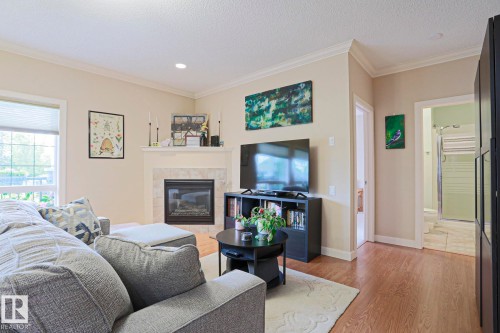 Living area featuring a fireplace with a stone surround and mantel, light wood-finish flooring, a window with blinds, recessed lighting, and crown molding - 13 7293 South Terwillegar Drive, Edmonton, AB 
