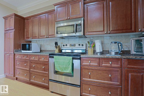 Expansive kitchen featuring wood-finish cabinetry, stainless steel appliances including a range and microwave, granite-look countertops, and a neutral tile backsplash - 13 7293 South Terwillegar Drive, Edmonton, AB 
