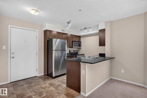 Open-concept kitchen featuring dark wood cabinetry, stainless steel appliances, a black countertop breakfast bar, and track lighting - 205 11816 22 Avenue, Edmonton, AB 