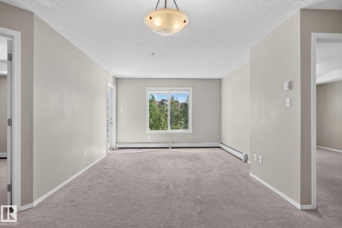 Open living area featuring neutral carpeting, light-toned walls, and a large window with exterior foliage - 205 11816 22 Avenue, Edmonton, AB 