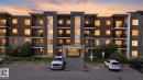 Multi-story building exterior featuring stone and wood-finish siding, multiple private balconies with black railings, a paved parking lot, and mature landscaping - 205 11816 22 Avenue, Edmonton, AB 
