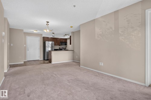 Open concept living area with carpeting, featuring a kitchen with dark wood cabinetry, stainless steel appliances, and a breakfast bar - 205 11816 22 Avenue, Edmonton, AB 