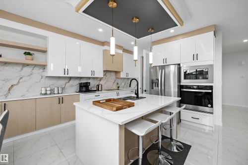 Contemporary kitchen featuring a central island with a white countertop, two-tone cabinetry, integrated stainless steel appliances, a complementary backsplash, and pendant lighting - 403 Pine Point, Leduc, AB 