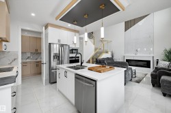 Open-concept living area featuring a kitchen island with integrated sink, stainless steel appliances, a modern fireplace with a tiled surround, and a staircase with glass railings - 