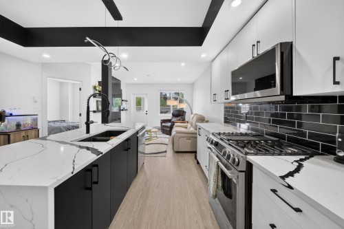 Modern kitchen featuring an expansive island with a large sink, white countertops with prominent veining, matte black cabinetry, stainless steel appliances, and a black subway tile backsplash - 626 Astoria Way, Devon, AB 