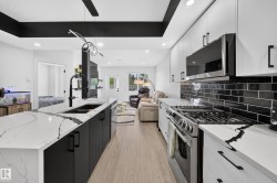 Modern kitchen featuring an expansive island with a large sink, white countertops with prominent veining, matte black cabinetry, stainless steel appliances, and a black subway tile backsplash - 
