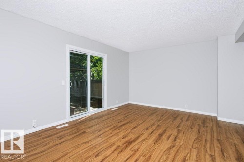 Spacious room featuring wood-finish flooring, light grey walls, and a white sliding glass door providing outdoor access - 24 14511 52 Street, Edmonton, AB 