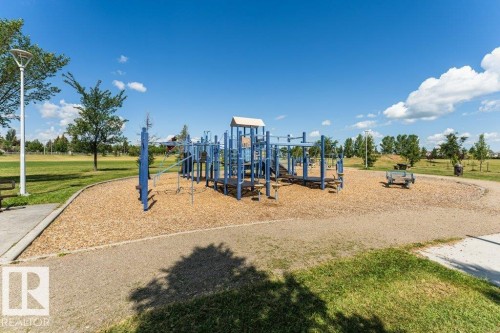 Community park featuring a blue multi-level playground structure on a wood chip surface, surrounded by expansive green spaces and mature trees - 24 14511 52 Street, Edmonton, AB 